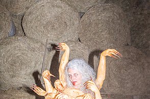 Foto von einer Person in einem Kostüm mit mehreren Armen die im Heu liegt