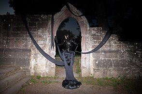 Person in schwarzem Kostüm steht vor einer Steinmauer und posiert. Teile des Kostüms sind lang genug um über der Mauer zuhängen