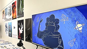 Foto eines Bildschirms auf dem Animationen laufen. Im Hintergrund hängen Poster und auf einem Tisch liegen Storyboards
