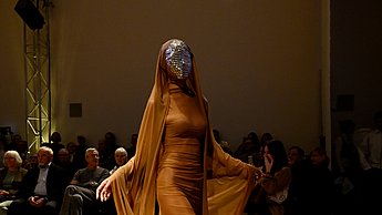 Model trägt ein orangenes Kostüm mit einer silbernen Maske