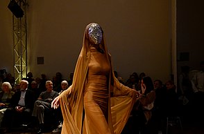 Model trägt ein orangenes Kostüm mit einer silbernen Maske