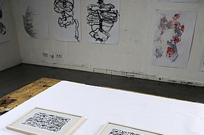 Vier gerahmte Gemälde liegen auf einem Tisch. Im Hintergrund sind weitere Bilder aufgehängt.