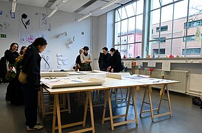 Foto eines Zimmers in welchem ein großer Tisch mit Zeichnungen steht. Menschen betrachten diese.