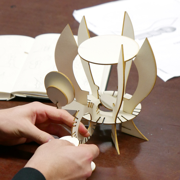 Nahaufnahme wie ein Modell aus gefrästen Elementen zusammengebaut wird, im Hintergrund Skizzen davon.