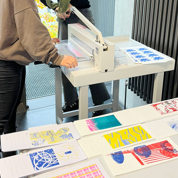 Aufnahme, wie Studierende ihre grafischen Werke auslegen und an einer Schneidemaschine zurechtschneiden.